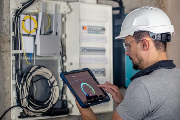 Électricien équipé inspectant une armoire électrique de distribution et de télécommunications dans un bâtiment industriel. Il utilise une tablette pour des mesures précises et complexes, qui pourraient s'apparenter aux calculs précis effectués par un compteur Geiger.
